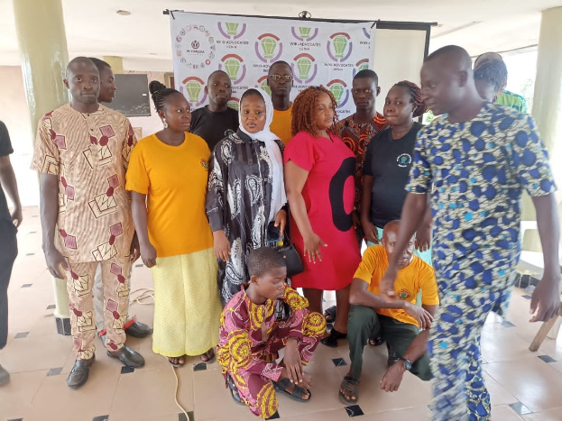 Eine Gruppe von Menschen steht vor einem Banner mit der Aufschrift "Advocates in Ghana: Ein Schritt-für-Schritt-Führer" mit Stühlen, Säulen, einem Fernseher und Deckenleuchten im Hintergrund.