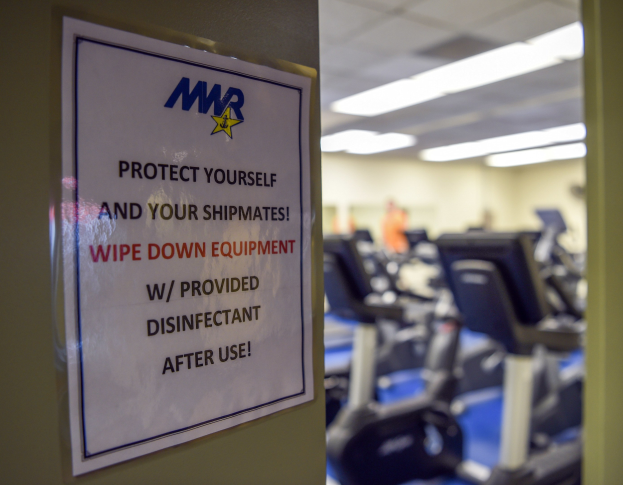 Zeichen an einer Turnhalle, auf dem steht: "Schütze Dich und Deine Schiffskameraden" mit Laufbändern und Deckenleuchten im Hintergrund.