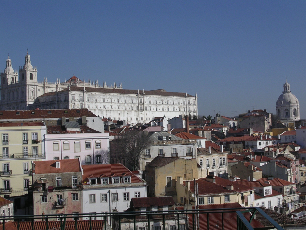 Blick auf Lissabon von einem Hügel aus, mit Gebäuden und Bäumen im Vordergrund und einem klaren blauen Himmel im Hintergrund.