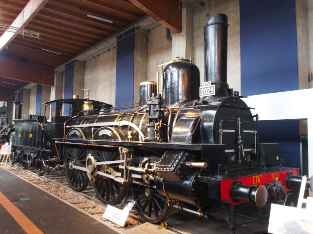 Alte Dampflokomotive in Museums Setting mit umliegenden Eisenbahnschienen, Informationsschildern und anderen Ausstellungen, vor einer Wand mit Deckenbeleuchtung.