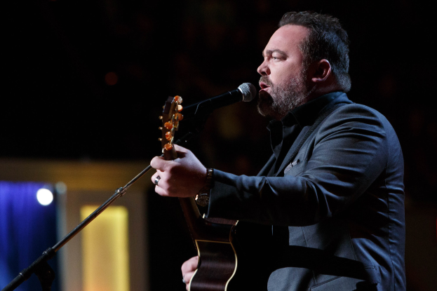 Ein Mann in einem Anzug singt in ein Mikrofon, während er eine Gitarre spielt, mit Dierks Bentley im Hintergrund, bei gedämpfter Beleuchtung.
