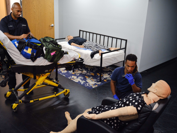 Eine Gruppe von Menschen in einem Raum mit einer Puppe auf einer Bahre, zwei Personen auf Betten liegend, eine Person stehend und ein Objekt haltend und eine weitere Person auf einem Stuhl sitzend, mit einer Tasche auf der Bahre und einer Matte auf dem Boden, vor einer Wand und einer Tür im Hintergrund.