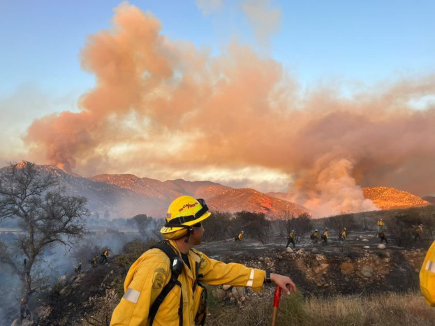 Ein Feuerwehrmann in einer gelben Jacke und einem Helm steht vor einer großen Rauchwolke, hält einen Stock und hat Bäume, Hügel und einen klaren blauen Himmel im Hintergrund.