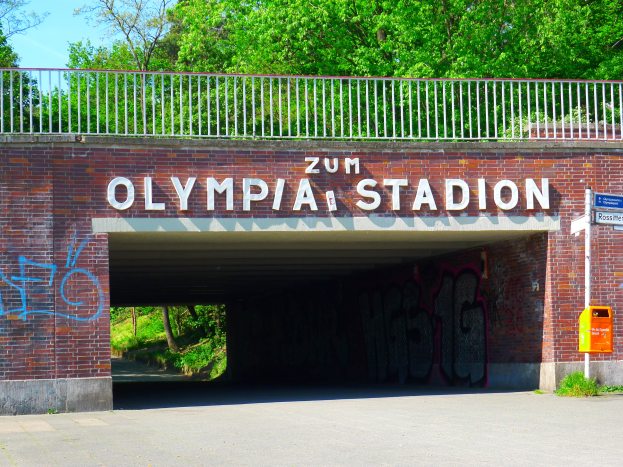 Der Eingang zum Olympiastadion in Berlin, Deutschland, mit einer Brücke mit Text, einem Metallzaun, einer Tafel, einer Kiste, Pflanzen, Gras, einer Gruppe von Bäumen und einem bewölkten Himmel.
