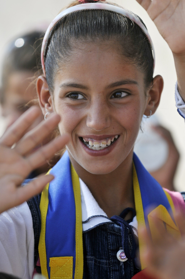 Ein junges Mädchen in Schuluniform lächelt und winkt in die Kamera, mit unscharfen Figuren im Hintergrund.