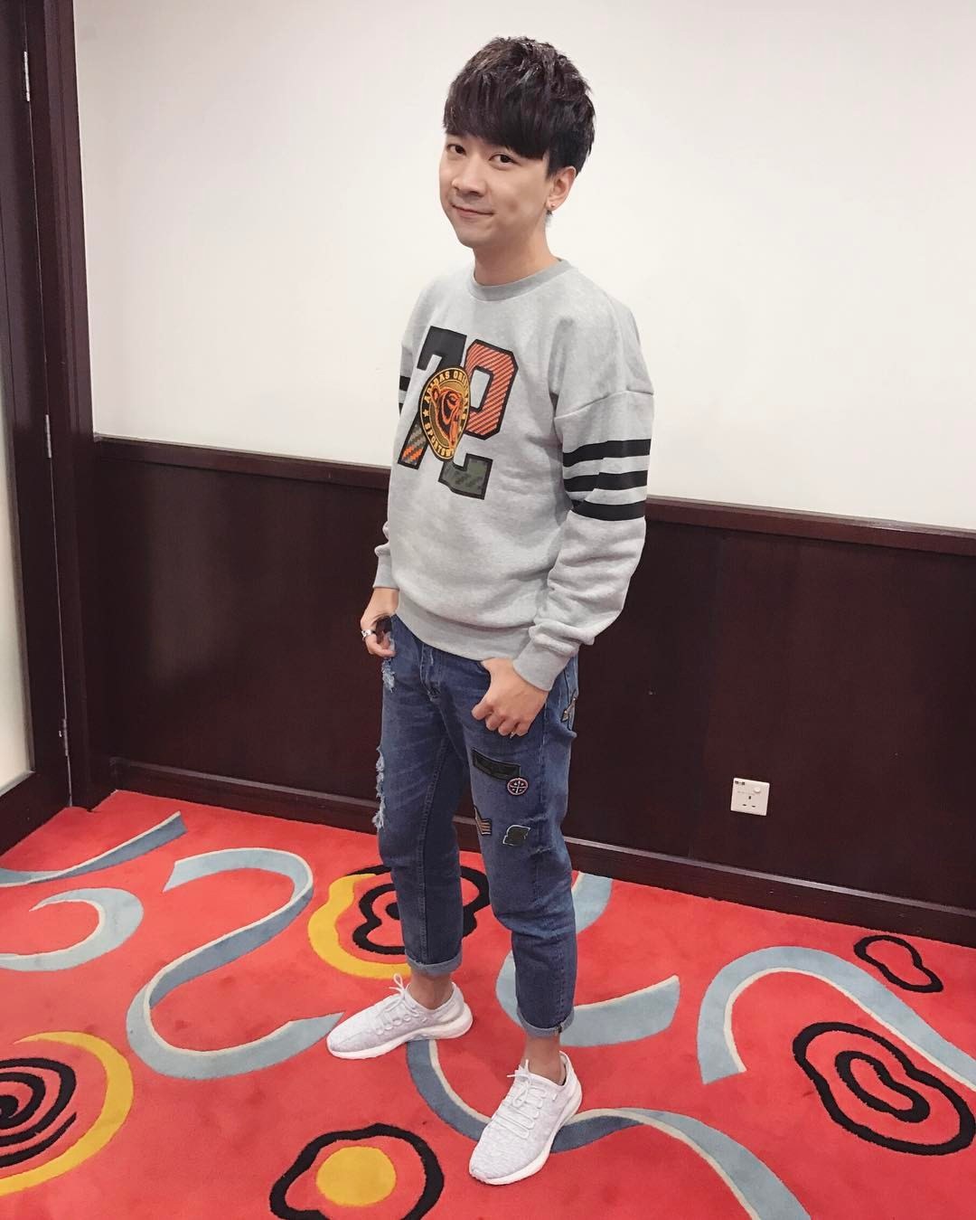 A young man in a grey sweatshirt and blue jeans stands smiling on a red carpet at a Korean fashion show, with a switchboard visible in the background.
