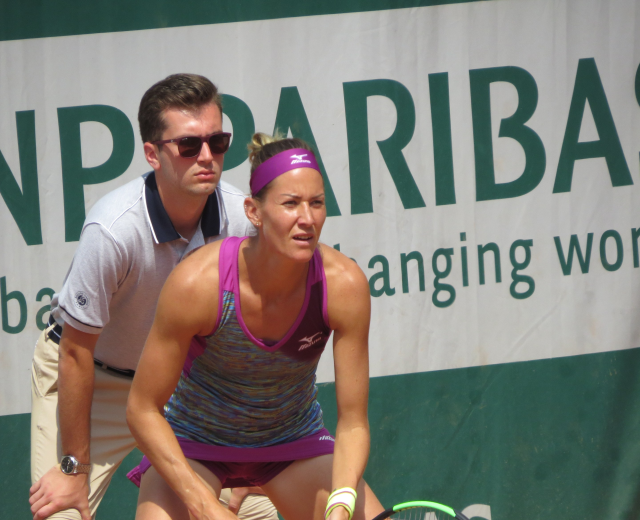 Zwei Personen spielen Tennis auf einem Court, wobei die Frau einen Schläger hält und der Mann eine Brille und eine Uhr trägt; im Hintergrund ist ein Banner mit Text zu sehen.