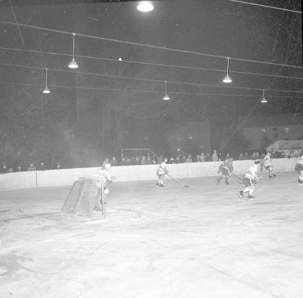 Schwarzes und weißes Bild einer Gruppe, die Hockey auf einem Eisplatz nachts spielt, mit einem Netz auf der linken Seite, Spielern mit Helmen und Stöcken und Zuschauern im Hintergrund.