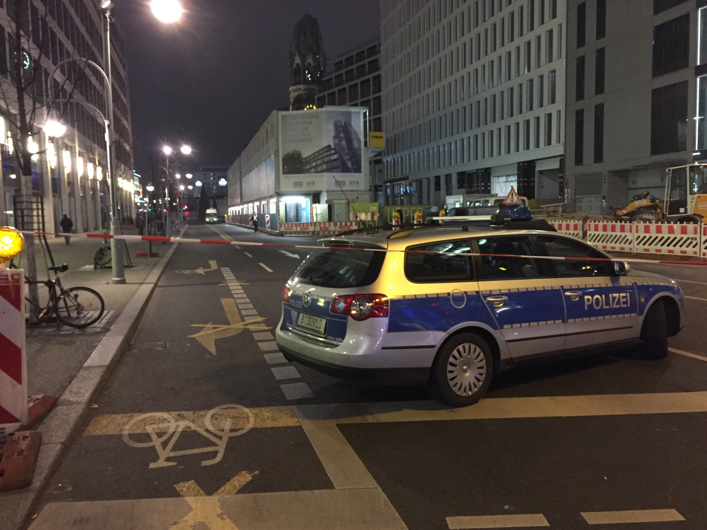 Ein Polizeiwagen auf einer Stadtstraße bei Nacht geparkt, umgeben von Gebäuden, Straßenmöbeln, Fahrzeugen und Menschen, mit einer Statue im Hintergrund.