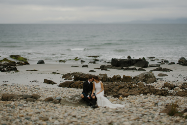 Braut und Bräutigam umarmend und küssend auf einem felsigen Strand mit dem Ozean und dem Himmel im Hintergrund.