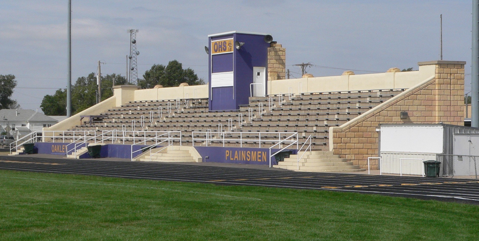 A football field with green grass, bleachers with railings, a wall with text, dustbins, utility poles with wires, trees, buildings with windows, and a cloudy sky.