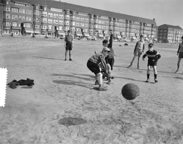 Schwarzes und weißes Bild von jungen Jungs, die auf einem Feld Fussball spielen, mit einem Ball in der Mitte, Gebäuden, Pfählen und einem klaren Himmel im Hintergrund.