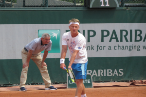Zwei Männer spielen Tennis auf einem Sandplatz, wobei einer in einem weißen T-Shirt und blauen Shorts einen Schläger hält, ein grünes Banner im Hintergrund und ein Zaun und eine Anzeigetafel im Hintergrund.