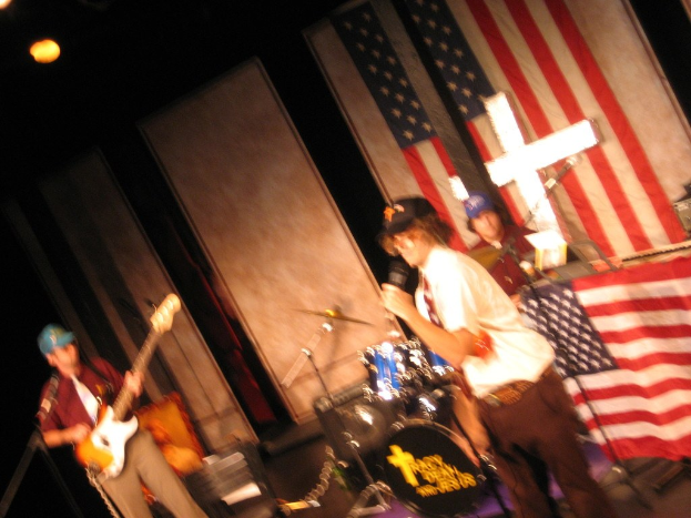 Gruppe von Menschen, die Musikinstrumente spielen, mit Equipment drumherum, amerikanischer Flagge im Hintergrund und einer Wand dahinter.