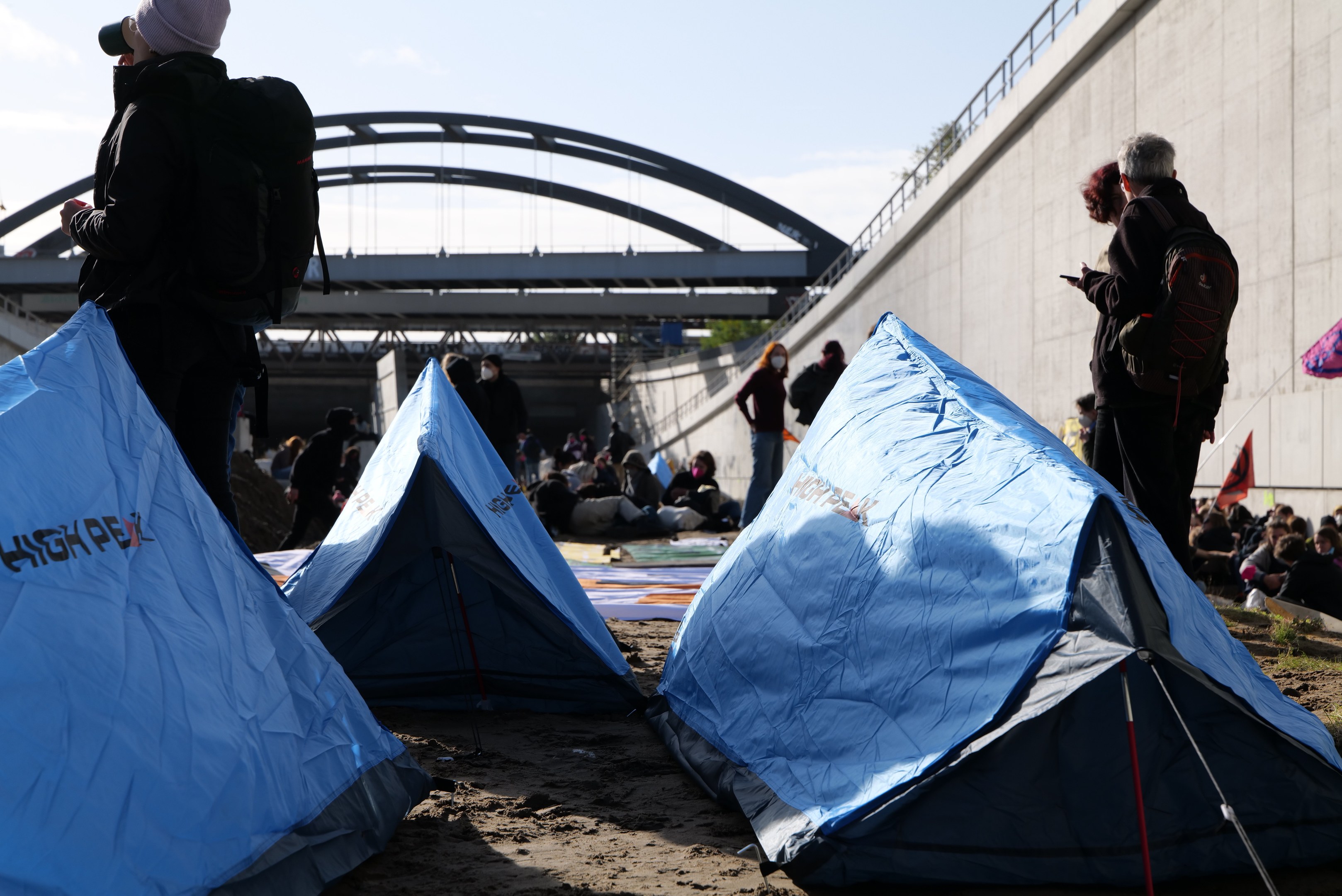 Eine Gruppe von Menschen sitzt auf einem sandigen Strand in der Nähe von Zelten, mit einer Wand und einer Brücke im Hintergrund, die an einer Klimawandel-Demonstration teilnehmen.