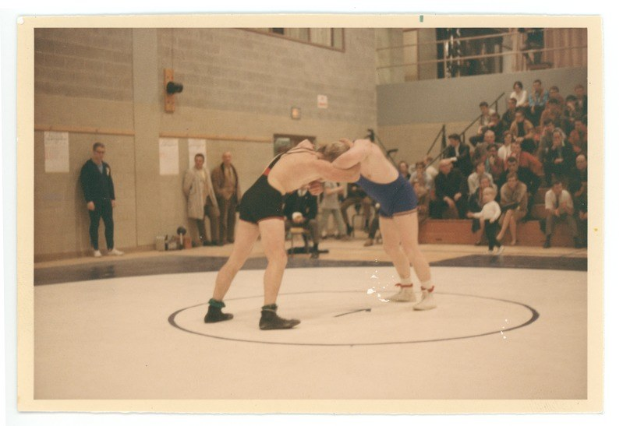 Zwei Wrestler kämpfen in der Mitte einer Menge, während Zuschauer aufmerksam zuschauen; eine Wand mit angeklebten Papieren bildet den Hintergrund.