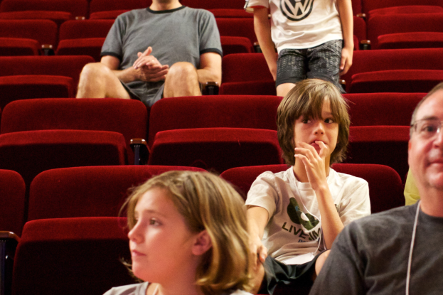 Eine Gruppe von Menschen sitzt in einem Kino mit roten Stühlen, während ein Junge in einem weißen T-Shirt und schwarzen Hosen in der Mitte steht, umgeben von anderen in verschiedenenfarbiger Kleidung.