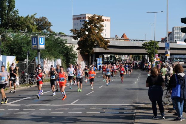 Gruppe von Menschen bei einem Marathon auf einer von Bäumen gesäumten Straße mit Fahrrädern, Schildern, einem Zaun, einer Brücke, Gebäuden und einem klaren blauen Himmel.