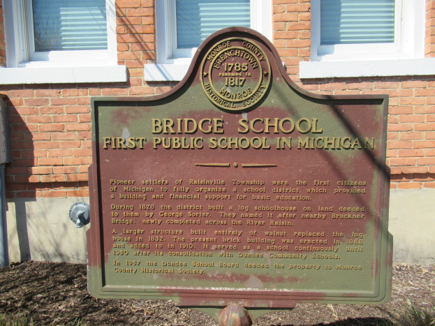 Ein Kreidetafel-Schild liegt auf dem Boden vor einem Backsteingebäude mit Fenstern und trägt die Aufschrift "Bridge School First Public School in Michigan."