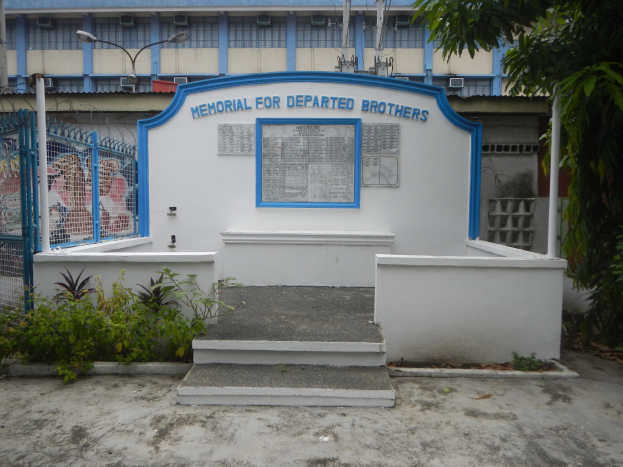 Gedenken mit Blumen und einem blauen-weißem Schild vor einem Gebäude bei bewölktem Himmel.
