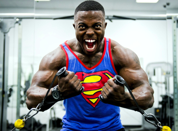 Ein Mann in einem Superman-Tanktop führt Klimmzüge in einem Fitnessstudio durch, umgeben von Fitnessgeräten unter Deckenbeleuchtung und trägt eine Uhr.
