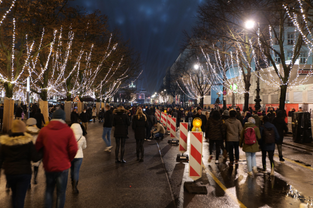 Eine Menschenmenge, die nachts eine Straße entlanggeht, beleuchtet von festlichen Weihnachtslichtern mit Bäumen und Gebäuden im Hintergrund.