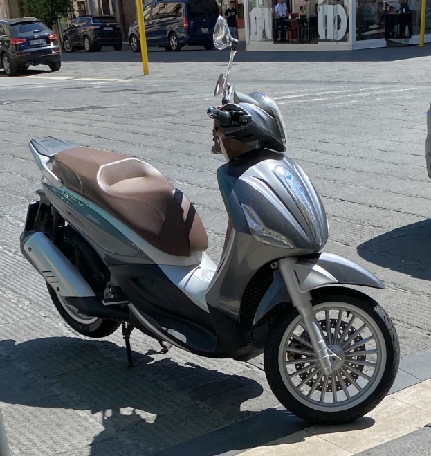 Ein Motorroller mit einem Helm darauf auf einer Straße geparkt, mit Gebäuden, Bäumen, Pfählen, Menschen und Gartenmöbeln im Hintergrund.