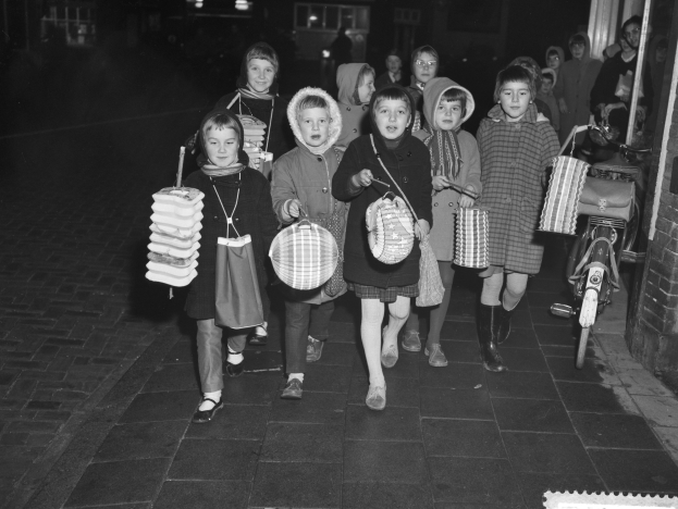 Gruppe von Kindern, die nachts eine Straße entlanggehen, jeder hält eine Tasche, mit einem Fahrrad an einer Wand gelehnt und Gebäuden im Hintergrund.