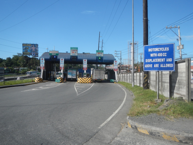Mautstation an einer Straße mit fahrenden Fahrzeugen, umgeben von Gras, Strommasten, einer Wand, einem Schuppen, Informationsschildern, Bäumen, entfernten Gebäuden und einem bewölkten Himmel.