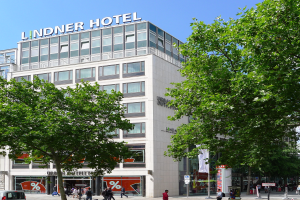 Das Lindner Hotel in Berlin, Deutschland, ein großes Glasgebäude mit Text auf der Oberseite, umgeben von Bäumen, Fahrzeugen auf der Straße und Menschen, die herumlaufen, mit dem Himmel im Hintergrund.