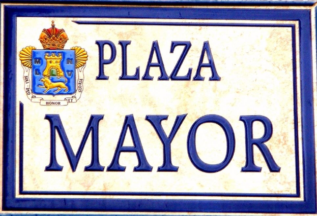 Rechteckiges weißes Schild mit schwarzer Schrift "Plaza Mayor" in fetter Schrift, blauer Rahmen und einem gekrönten Wappen-Logo an einer Gebäudefassade.