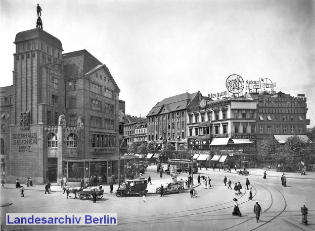 Schwarz-weiß-Foto einer belebten Berliner Citystraße mit Fußgängern, Fahrzeugen, einer Gebäudestatue, Beschriftungen, Pfosten, Bäumen und einem bewölkten Himmel.