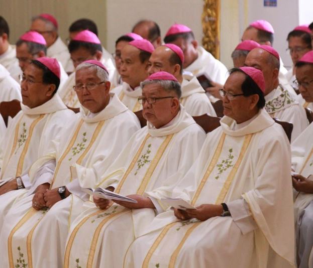 Eine Gruppe von Priestern in weißen Roben und pinken Mützen sitzt in einer Kirche und hält Bücher in der Hand, mit einer Wand im Hintergrund.