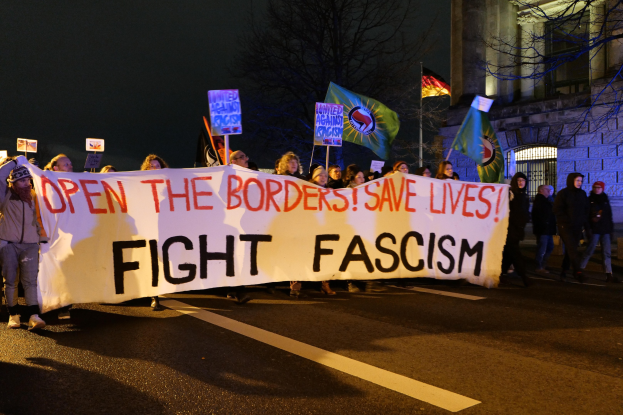 Eine Gruppe von Menschen marschiert auf einer von Bäumen gesäumten Straße, die eine Fahne mit der Aufschrift 'Öffne die Grenzen, rette Leben, kämpfe gegen Faschismus' trägt, mit einem Gebäude im Hintergrund.