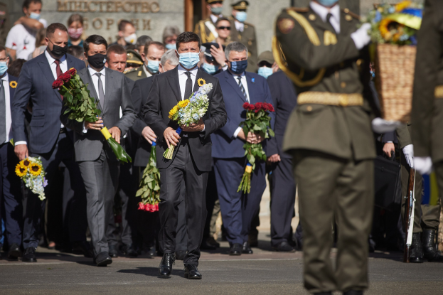 Gruppe von maskierten Männern in Anzügen mit Blumensträußen, mit einem uniformierten Wachmann mit einer Waffe auf der rechten Seite, vor einer Wand mit Gedenktext.