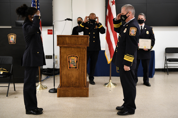 Gruppe uniformierter Personen auf einem Podium bei einer Vereidigung eines neuen Feuerwehrchefs, der eine Urkunde hält, mit Fahnen, Stühlen, Tischen und einer Wand im Hintergrund.