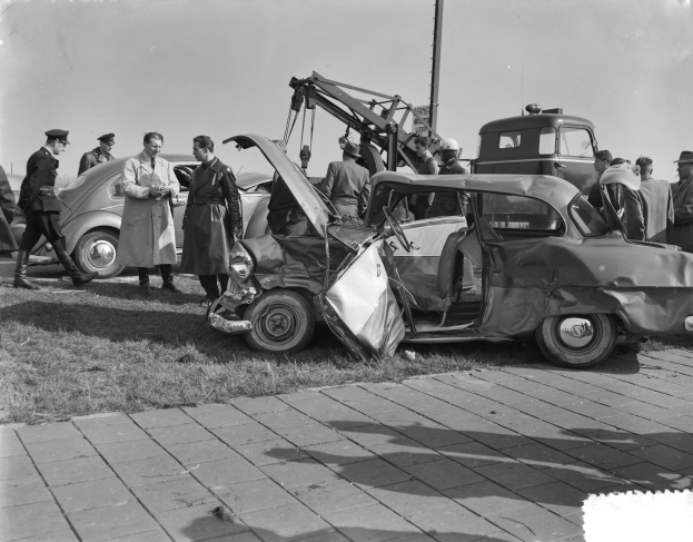 Schwarz-weißes Bild eines verunglückten Autos am Straßenrand mit mehreren Menschen in der Nähe und einem Mast im Hintergrund, Gras bedeckt den Boden.