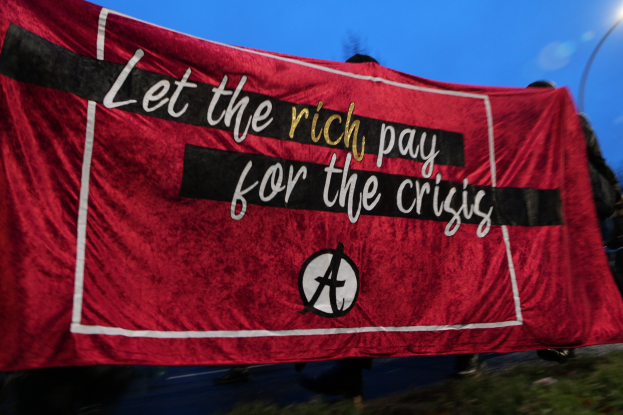 Eine Gruppe von Menschen marschiert auf einer Straße, die eine rote Fahne mit der Aufschrift "Lasst die Reichen für die Krise bezahlen" trägt, mit Bäumen, einem Laternenpfahl und einem klaren blauen Himmel im Hintergrund.