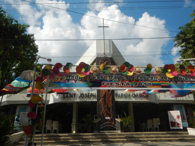Ein Gebäude mit einer "Fröhlichen Feiertagsgrüße St. Joseph der Arbeiter"-Schilder, Treppe zum Eingang, ein Torbogen mit Dekoration, Außenbestuhlung, Topfpflanzen, Bäume und ein bewölkter Himmel.