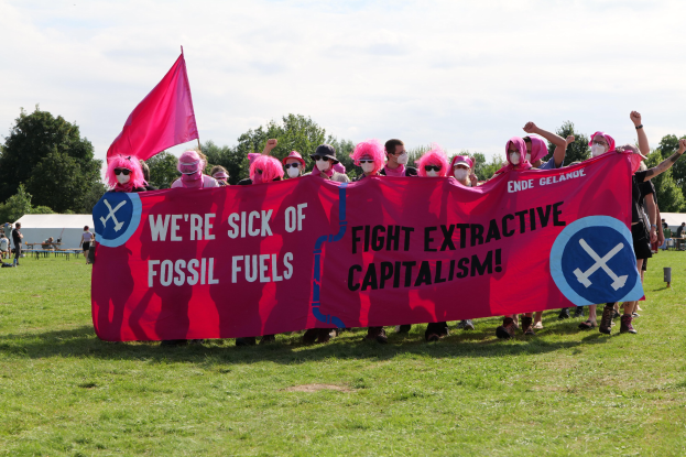 Gruppe von Menschen mit einem pinken Banner, auf dem 'Wir haben genug von fossilen Brennstoffen, kämpft gegen den extraktiven Kapitalismus' steht, auf einer grünen Wiese mit Zelten und Bäumen im Hintergrund unter einem klaren blauen Himmel.