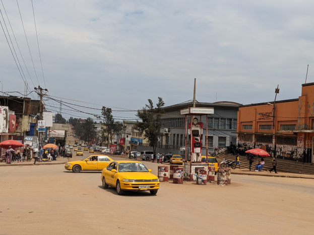 Ein gelbes Taxi fährt auf einer belebten Stadtstraße mit Gebäuden an den Seiten, während Fußgänger mit Regenschirmen auf dem Gehweg laufen und sich Wolken am Himmel befinden.
