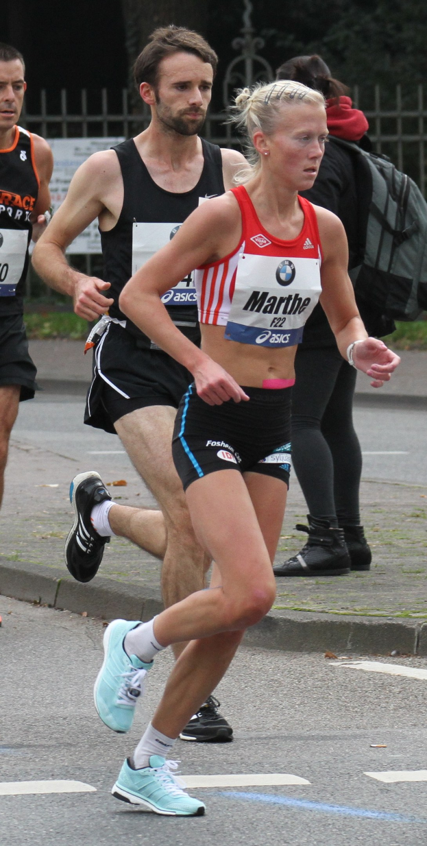 Eine Gruppe von Läufern bei einem Marathon, mit einer Frau in einem Sportdress, die an einem Zaun, Bäumen und Gras vorbeiläuft.