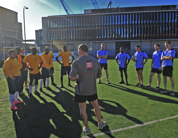 Gruppe von Männern auf einem Fußballfeld während eines Trainings, einer hält den Ball, mit Gebäuden, Laternenmasten und Kränen im Hintergrund.