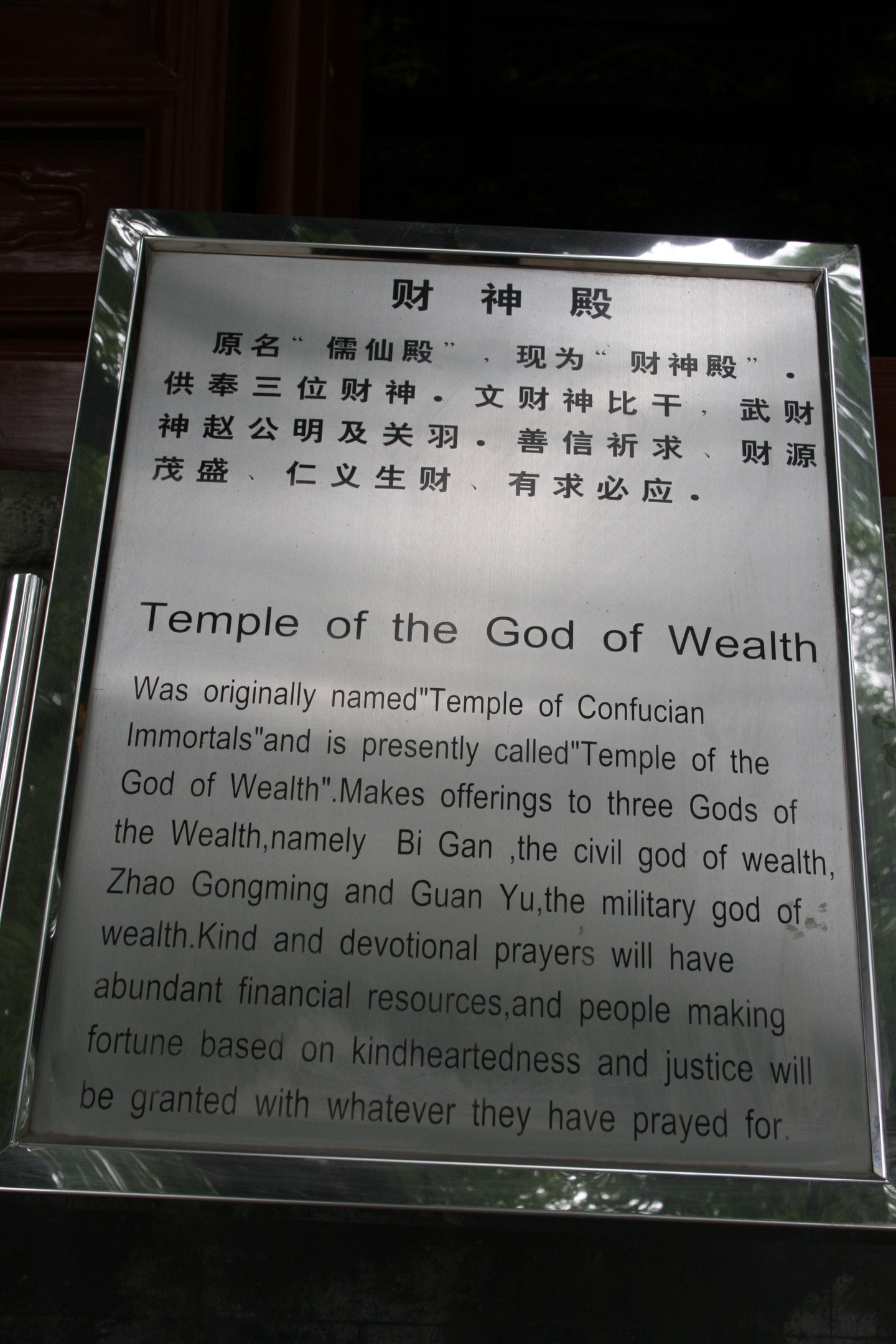 Ein Tempel, der dem Gott des Reichtums gewidmet ist, mit einer Namensplakette im Vordergrund und einer T├╝r im Hintergrund, umgeben von üppiger Vegetation.