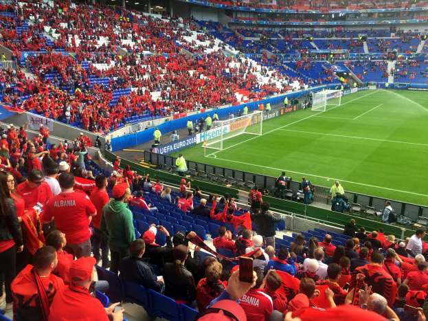 Eine große Menge Menschen in einem Stadion schaut ein Fußballspiel, mit Sitzplätzen, Fahnen, einem Torpfosten und Deckenleuchten.
