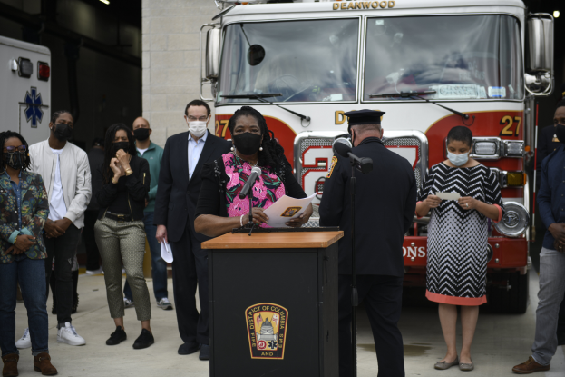 Bürgermeisterin von Chicago Lori Lightfoot steht an einem Rednerpult mit Mikrofonen und Papieren, spricht bei einer Pressekonferenz vor einem Feuerwehrauto, umgeben von maskierten Personen, mit einer beleuchteten Wand im Hintergrund.