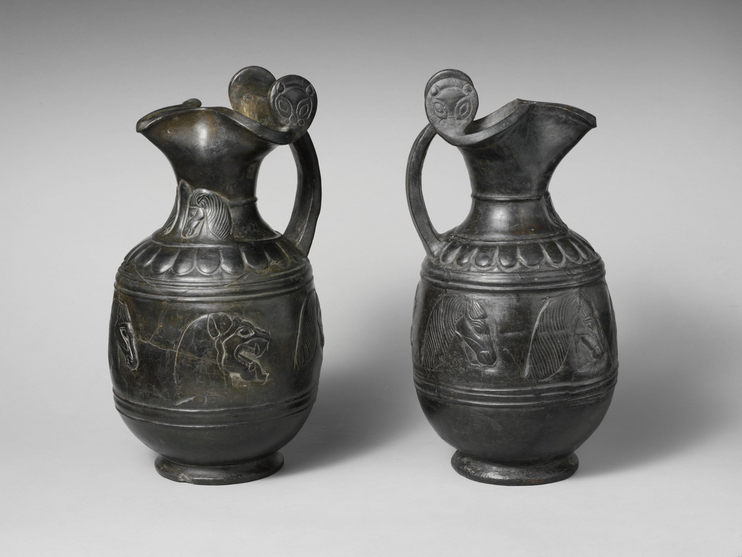 Two bronze jugs with glossy finishes, curved handles, long slender spouts, and intricate designs featuring geometric shapes and floral motifs.