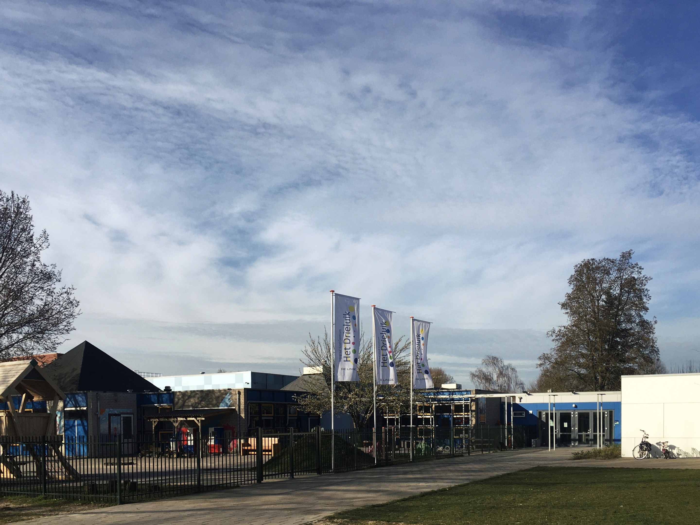 Gro├če Geb├Ąude mit einem grasigen Bereich davor, umgeben von einem Metallzaun, B├Ąumen, Bannern, einem geparkten Fahrrad, n├Ąheren H├Ąusern und einem bew├Âlktem Himmel, identifiziert als neuer Spielbereich für Kinder an einer Schule.