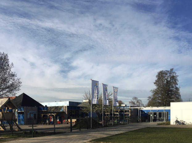 Gro├če Geb├Ąude mit einem grasigen Bereich davor, umgeben von einem Metallzaun, B├Ąumen, Bannern, einem geparkten Fahrrad, n├Ąheren H├Ąusern und einem bew├Âlktem Himmel, identifiziert als neuer Spielbereich für Kinder an einer Schule.