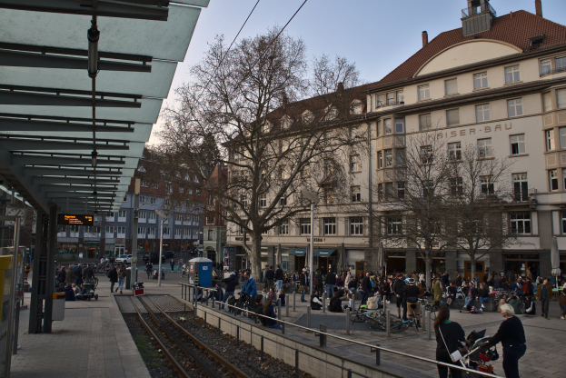 Eine belebte Stadtstraße mit einem Bahnhof auf der linken Seite, umgeben von Gebäuden, Bäumen und Laternen, wo Menschen gehen, auf Bänken sitzen oder stehen, mit Texttafeln und einem sichtbaren Himmel im Hintergrund.